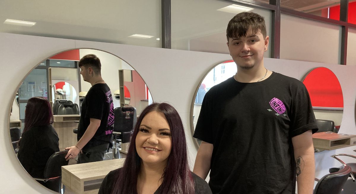 A female with long dark hair seated  with a young male with short dark hair standing behind in a hair salon with three large round mirrors in background.